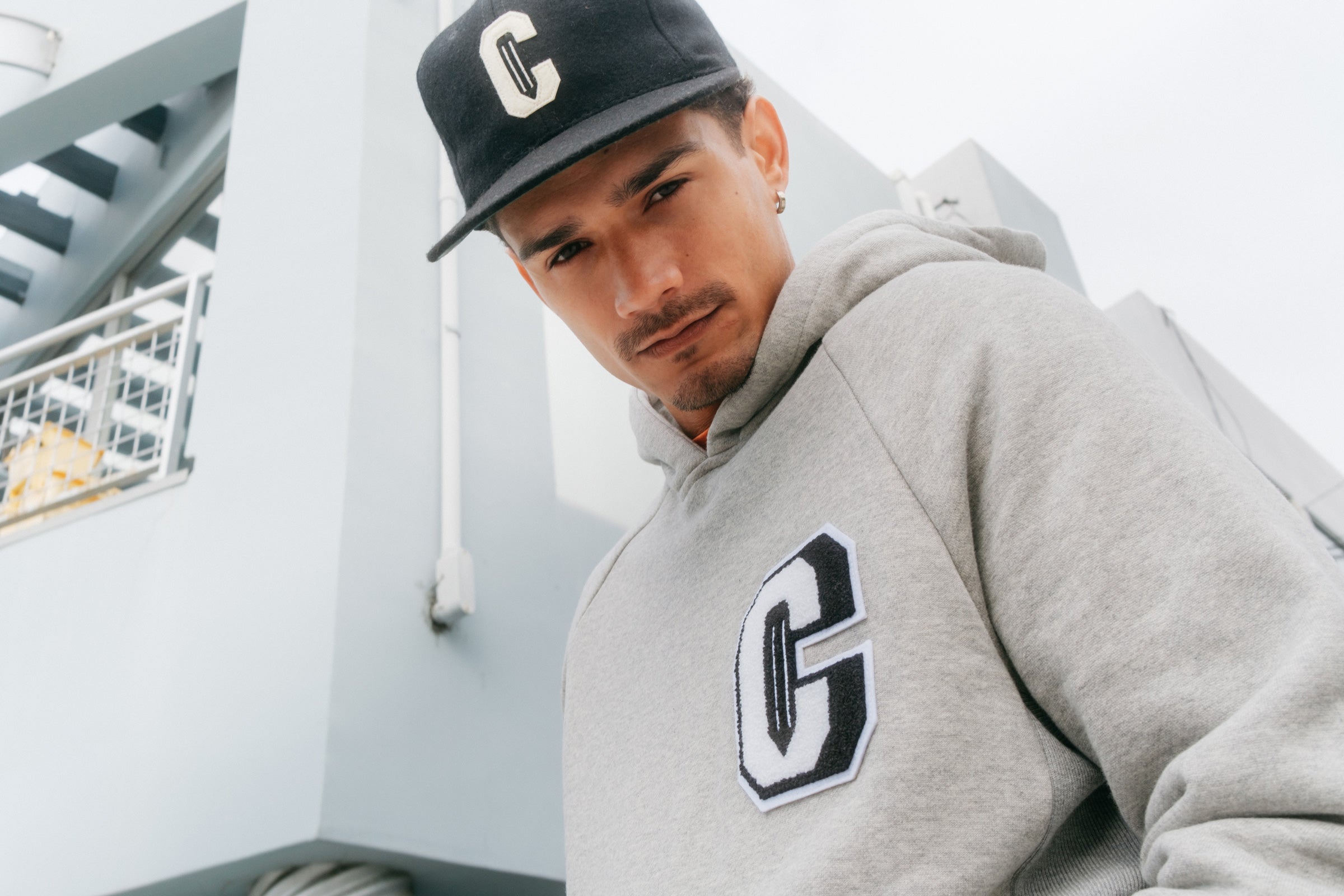 Model wearing the Varsity C Hoodie in grey and the Ebbets Wool C Cap in black and standing in front of a building.