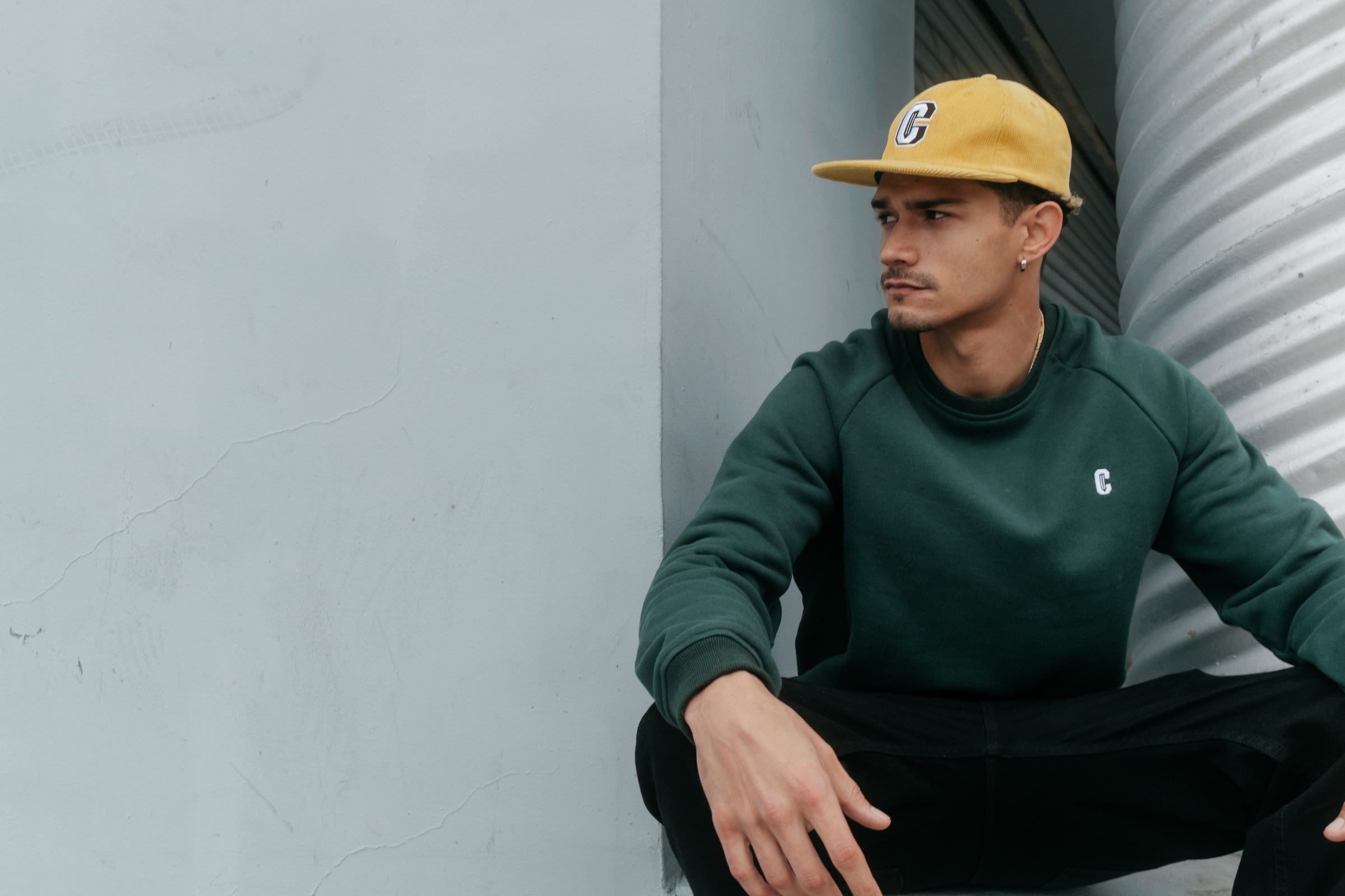 Model wearing the Varsity C Corduroy Cap in yellow and Icon Crewneck Sweatshirt in forest green and sitting at the corner of a building.