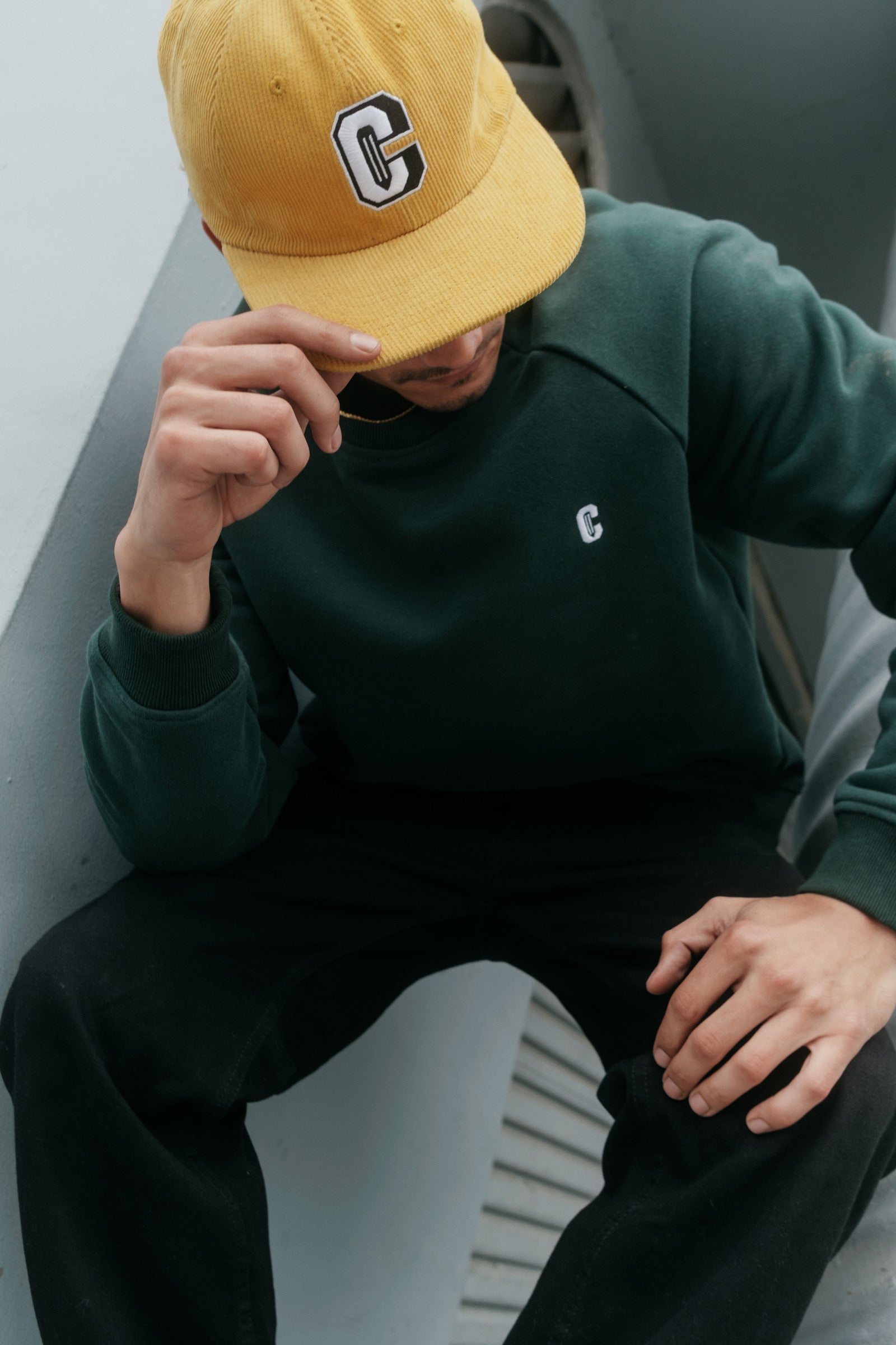 Model wearing the Varsity C Corduroy Cap in yellow and Icon Crewneck Sweatshirt in forest green and sitting at the corner of a building.