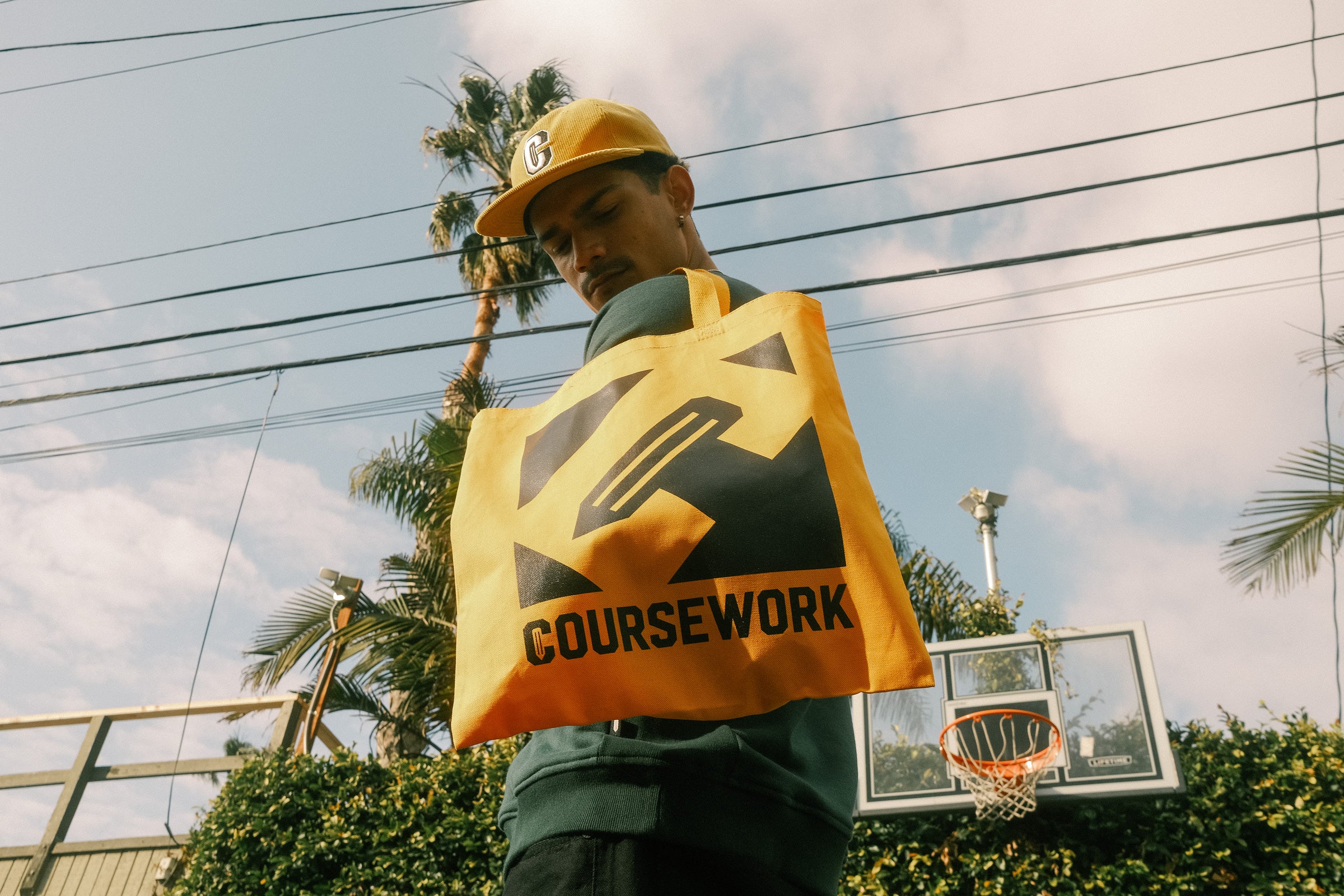 Model wearing the Varsity C Corduroy Cap in yellow and Icon Crewneck Sweatshirt in forest green holding a yellow tote bag..