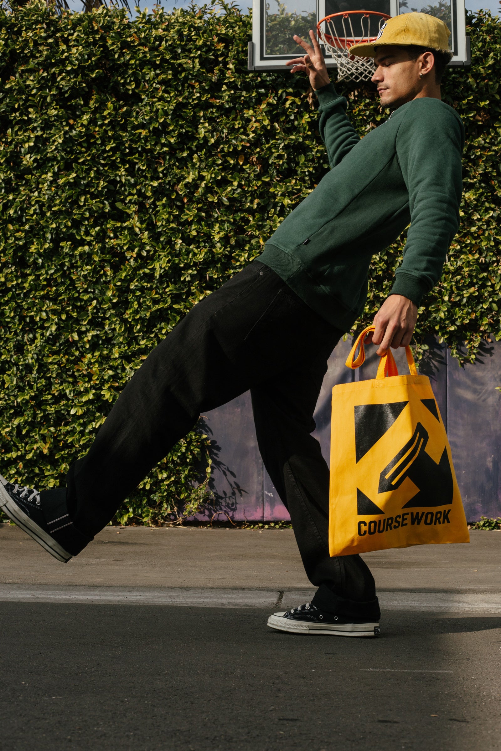 Model wearing the Varsity C Corduroy Cap in yellow and Icon Crewneck Sweatshirt in forest green and posing in front a basketball hoop and foliage..