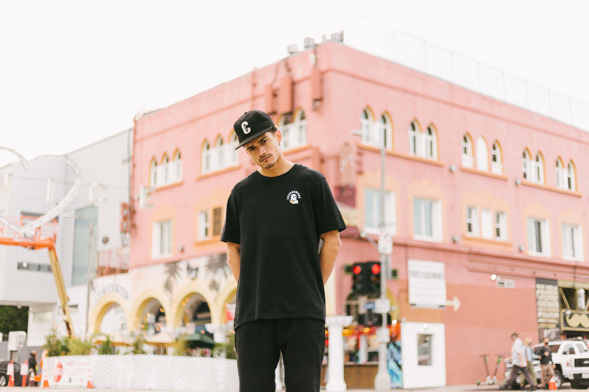 Model wearing the Ebbets Wool C Cap in black and the Required Reading Tee in black while standing on the sidewalk of an intersection.