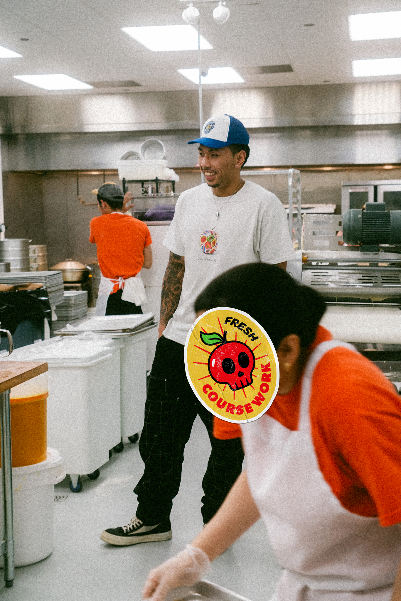 Model stands in kitchen of bakery with worker who accidentally gets into the image and has a fruit sticker design covering her face.