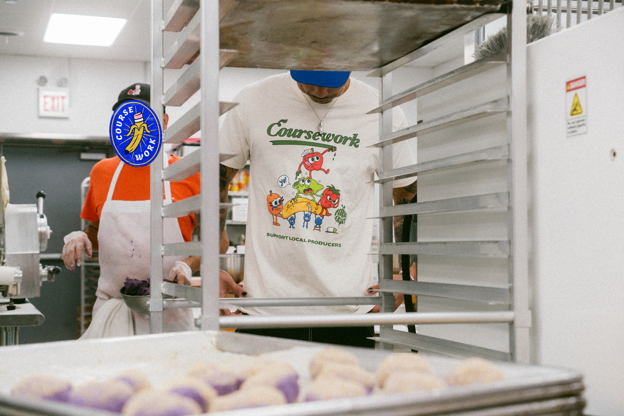 Model stands behind a cart in the kitchen of a bakery with the front of the t-shirt graphic in view within the cart