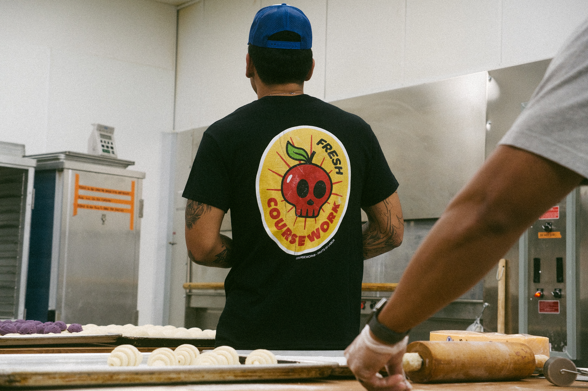 Model stands in bakery with back facing the camera and wears a t-shirt with a fruit sticker of an apple that is a skull