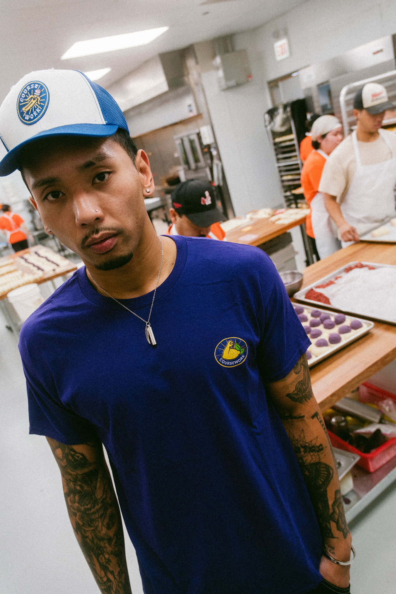 Model stands in kitchen of bakery with workers behind him making pateries