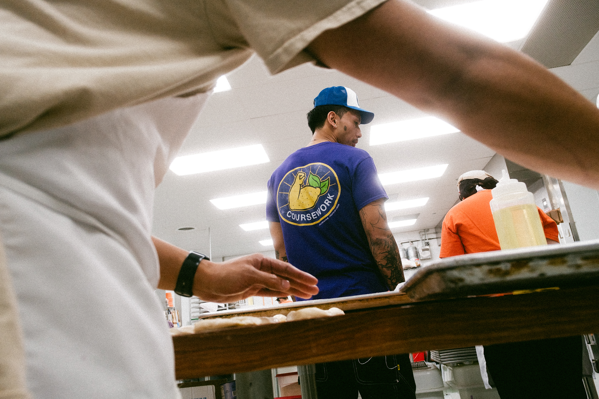 Model stands in the background of a kitchen in a bakery being viewed through the under arm of the worker making pasteries