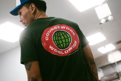 Model stands backside facing camera showing t-shirt graphic of fruit sticker of a watermelon that says Coursework Worldwide