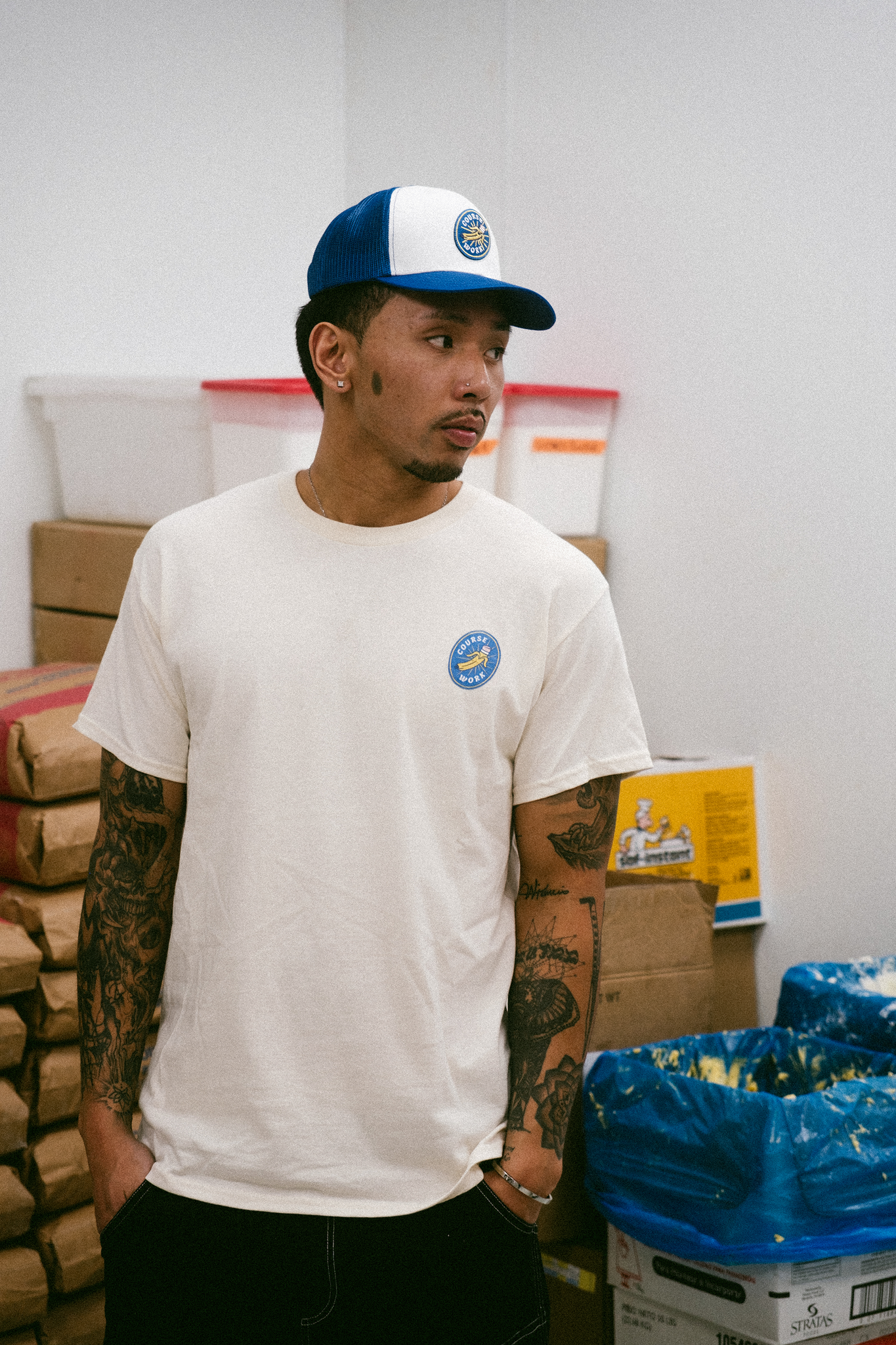 Model stands in kitchen of grocery store looking toward the right side of the image and wears a graphic tee and a trucker cap