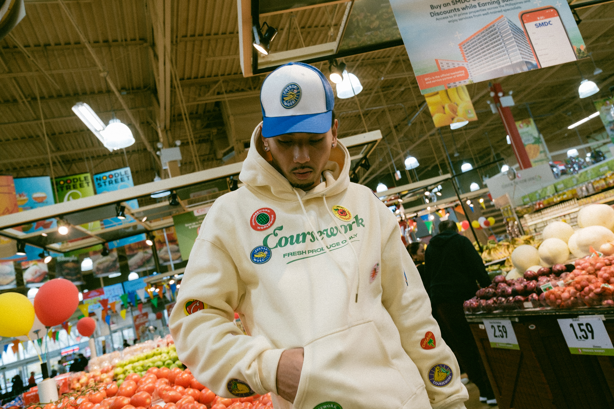 Model posing in hoodie and trucker cap in the produce section of the grocery store.