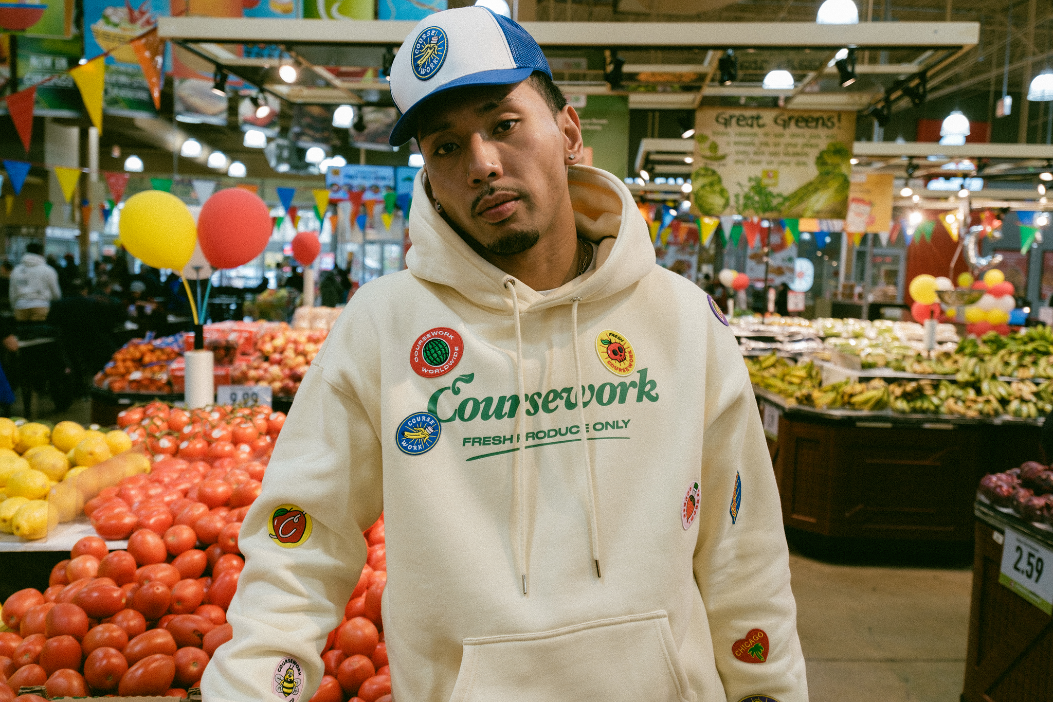 Model standing in hoodie and trucker cap in produce section of grocery store