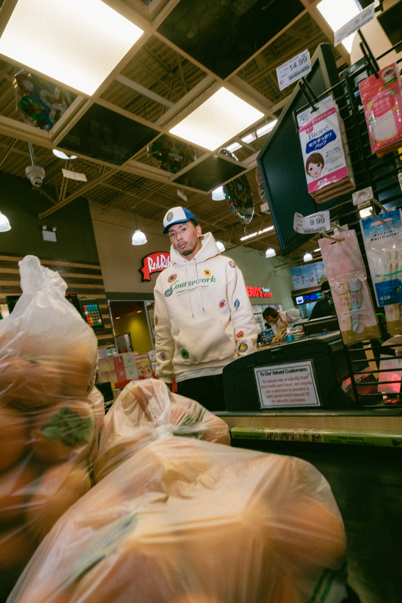 Model standing in grocery store with checkout lane and fruit in foreground.